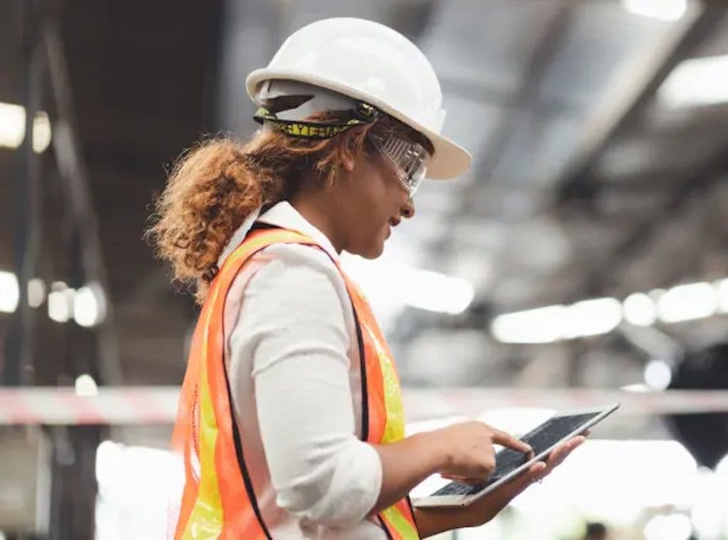 a woman wearing a hard hat and safety vest is using a tablet in a factory .