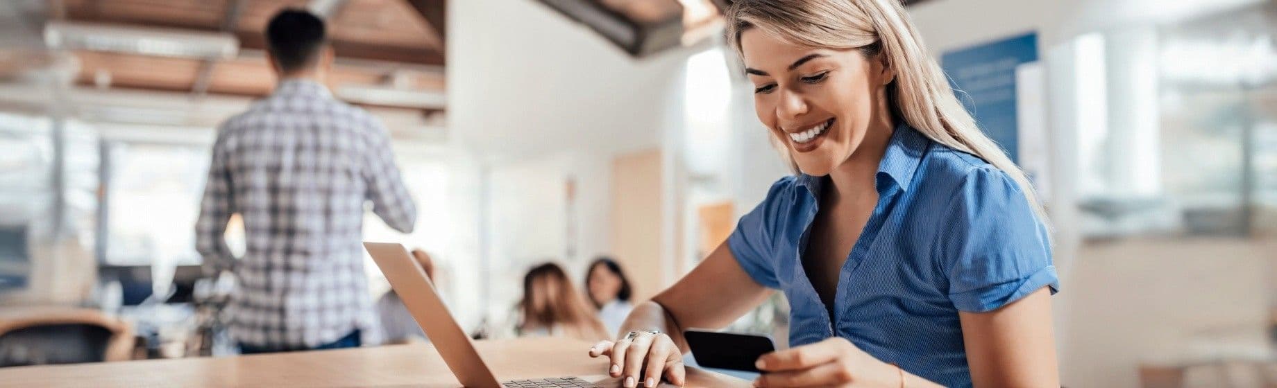 A smiling woman holding a credit card looks at a laptop in an office.