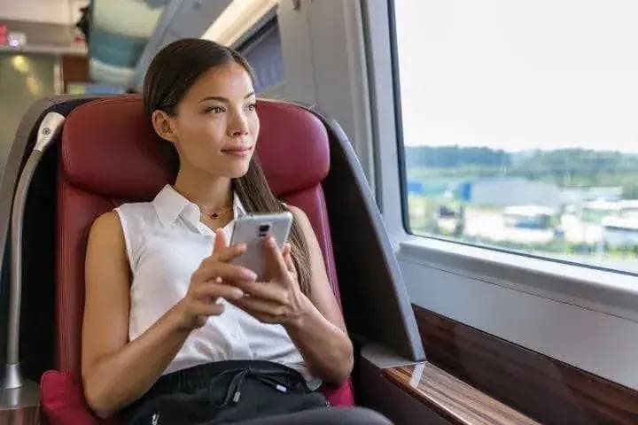 a woman is sitting on a train looking out the window while using her cell phone .