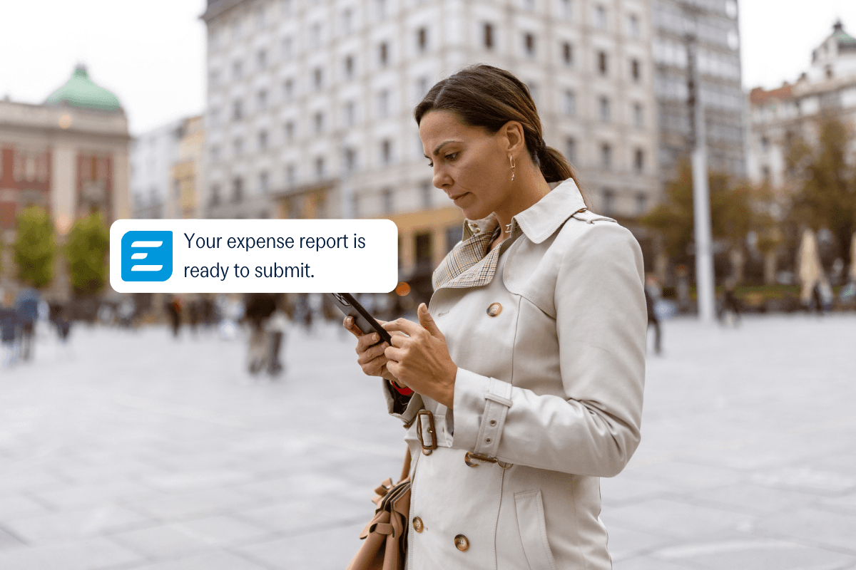 Woman in a city looking at her phone, which displays a notification: 'Your expense report is ready to submit.'