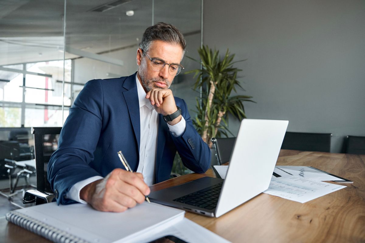 A man in a blue suit and glasses writes in a notebook at a desk with a laptop.