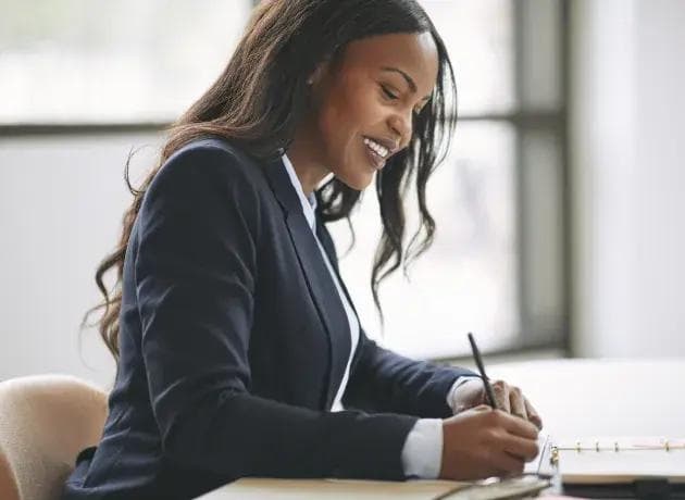 a woman in a suit is sitting at a desk writing on a piece of paper .