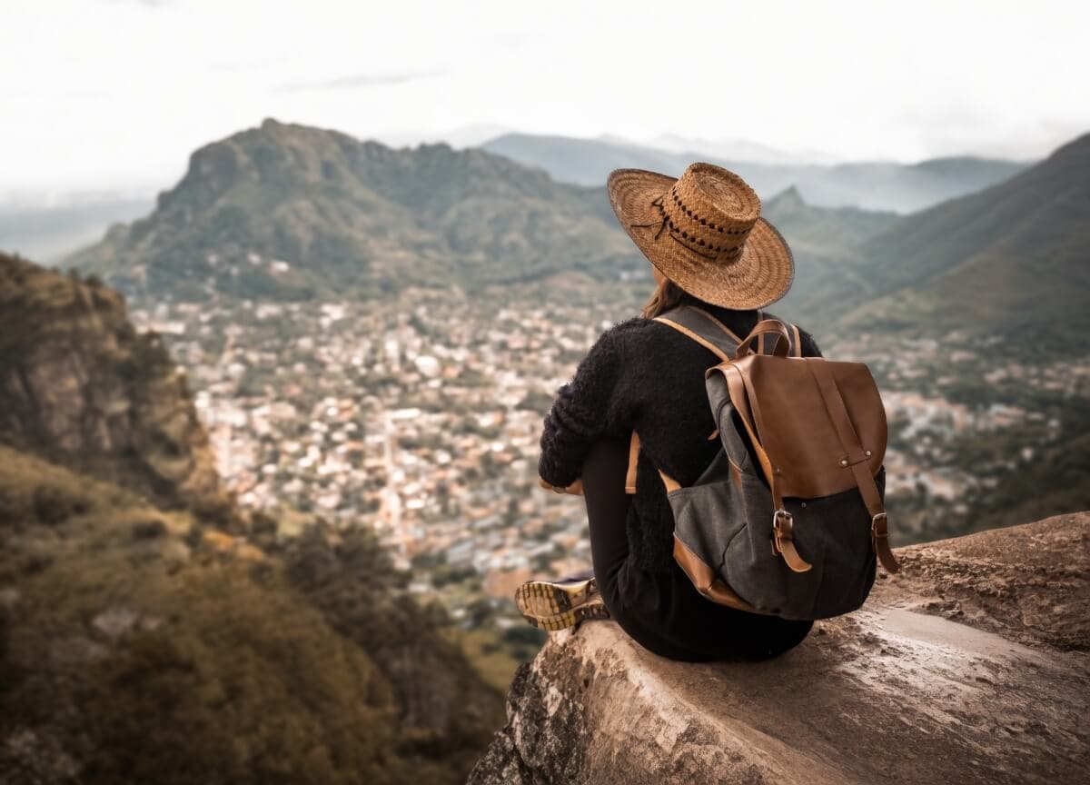 Person in a straw hat and backpack sitting on a cliff overlooking a town in a mountain valley.