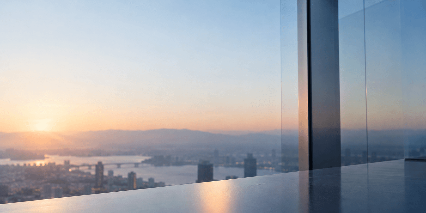 Sunset cityscape with a river and distant mountains, viewed from a high-rise window with reflections.