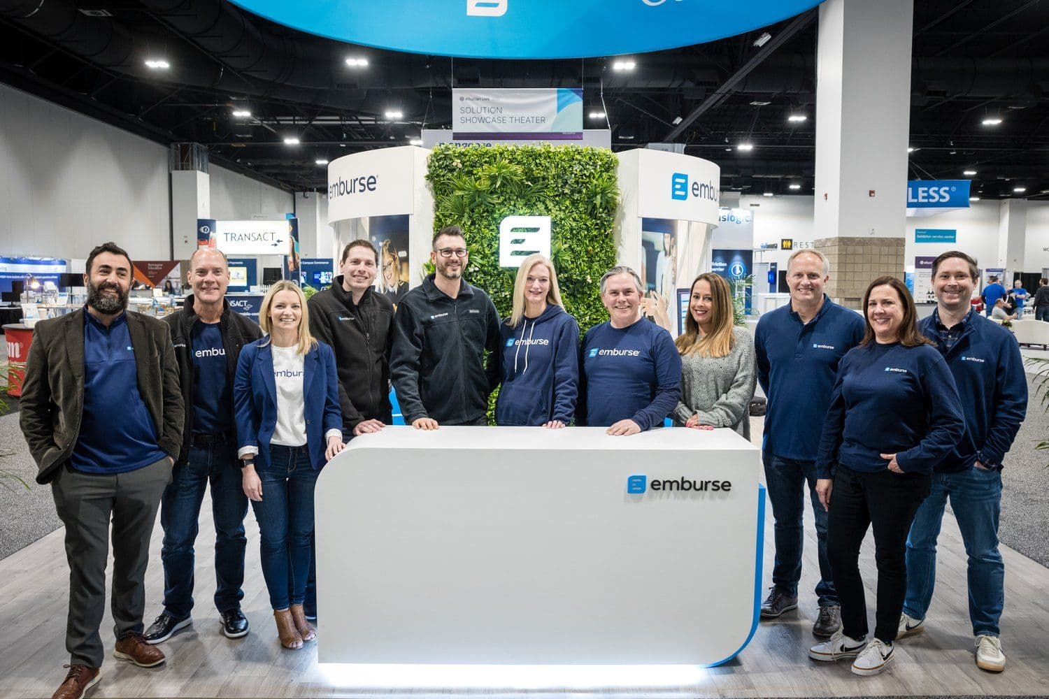 Twelve Emburse team members smile from their branded trade show booth with a prominent white counter and green wall.