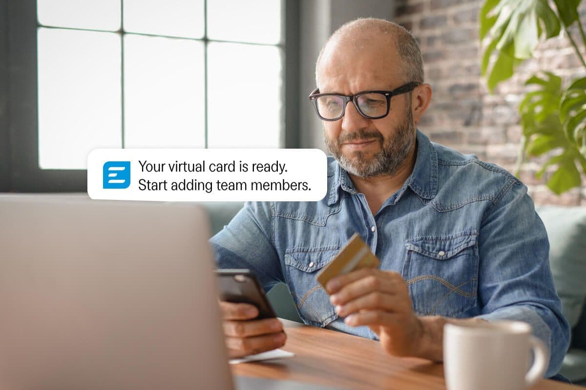 A man looks at his smartphone and a physical credit card, with an on-screen notification reading: "Your virtual card is ready. Start adding team members."
