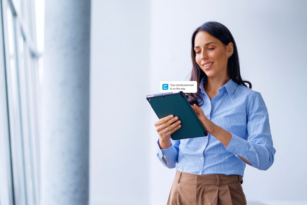 A smiling woman looks at a tablet displaying a notification: "Your reimbursement is on the way."